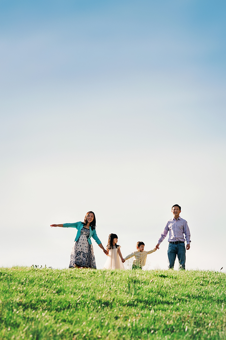 A happy family of four standing hand in hand at the top of the grassy slope.